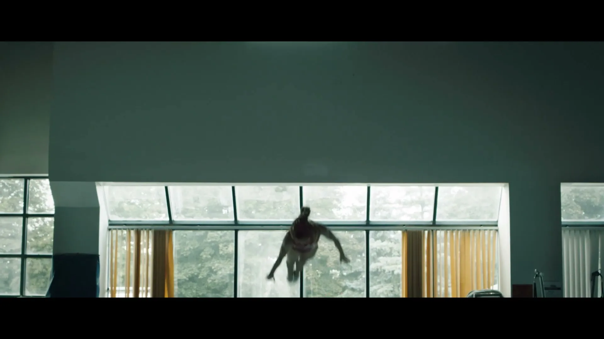 A female gymnast in a red leotard performs a flip in the air against a backdrop of large windows in a gym.