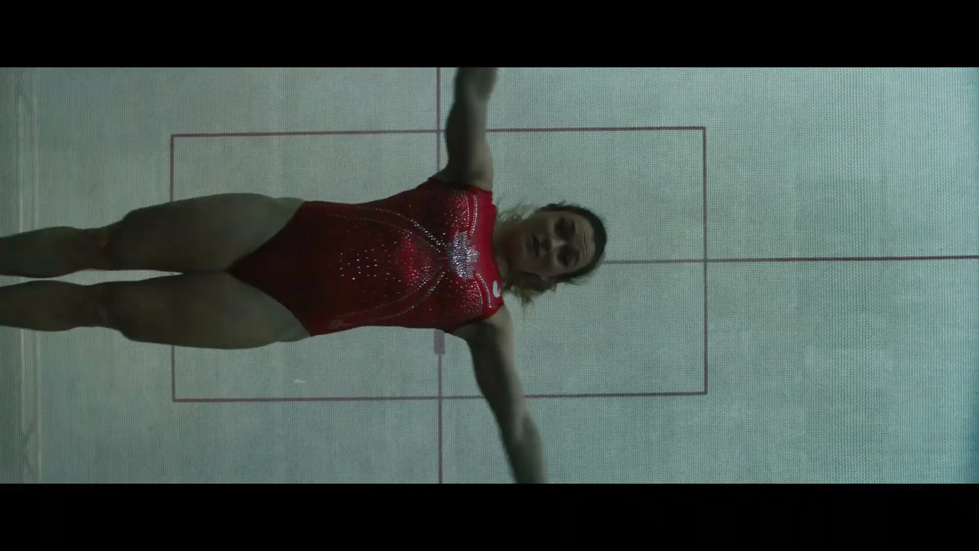 A female gymnast in a red leotard performs a routine on a mat, viewed from directly above in a gymnasium.