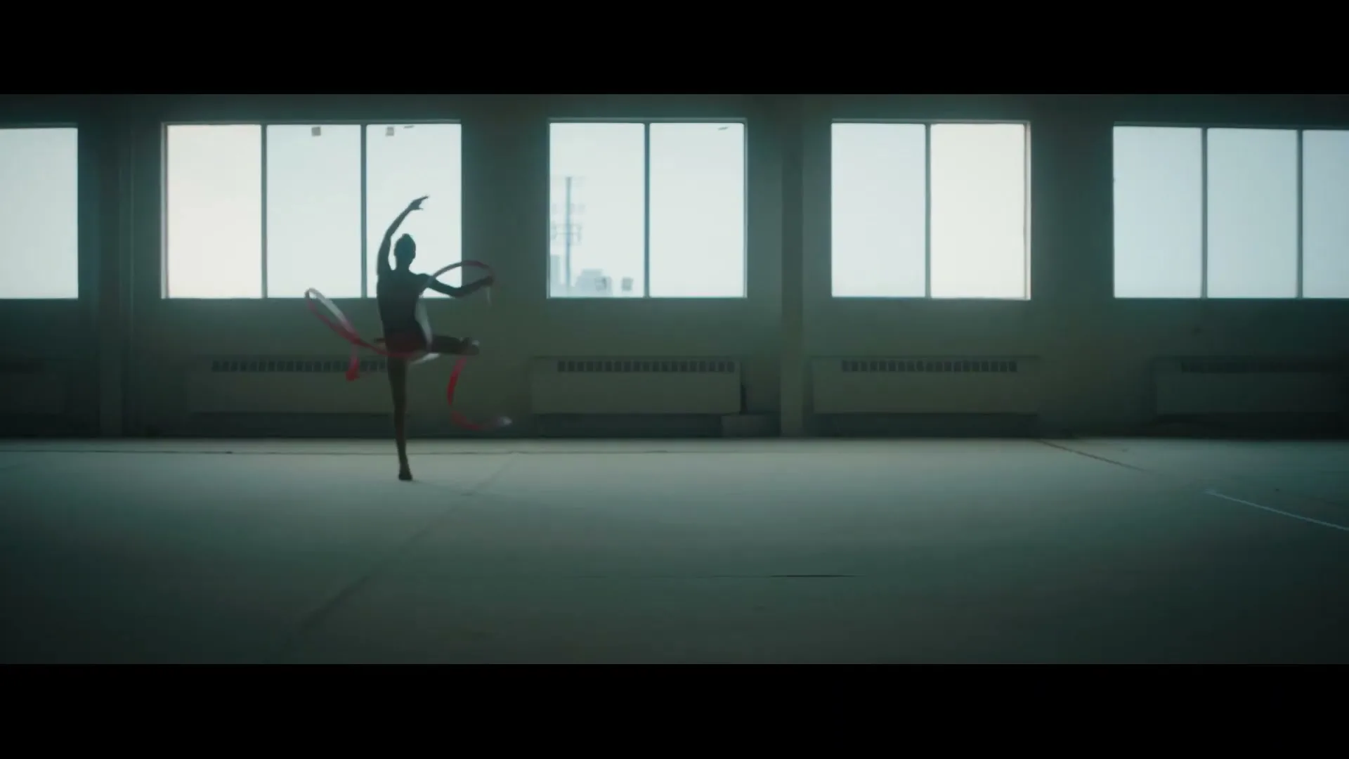 A female rhythmic gymnast in a leotard performs with a ribbon and hoop, silhouetted against large windows in a spacious gym.