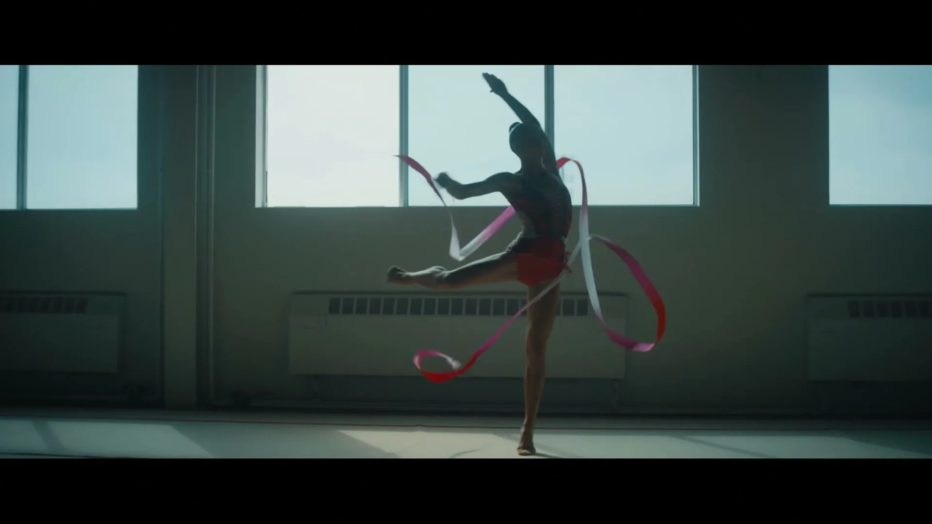 A female rhythmic gymnast in a red leotard performs a ribbon routine in a brightly lit gym with large windows.