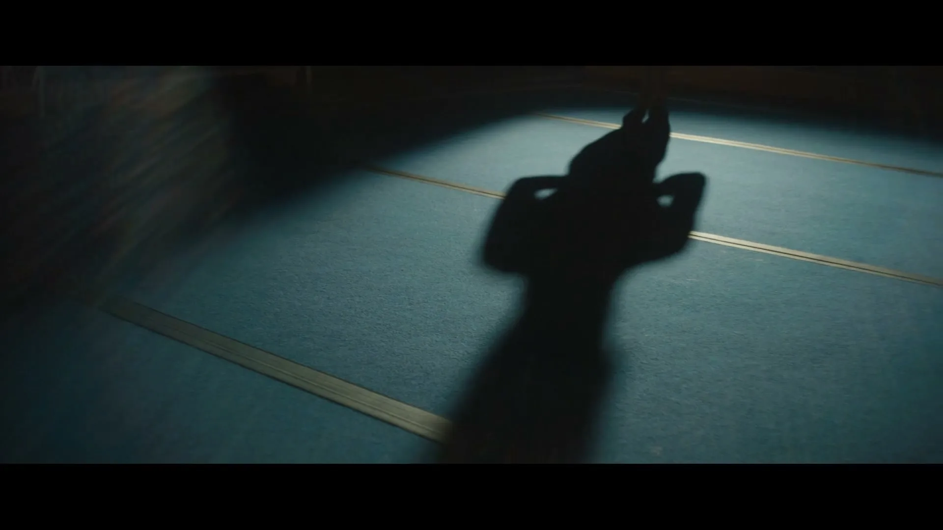 A gymnast's shadow is cast on a blue mat as they perform a handstand in a dimly lit gymnasium.
