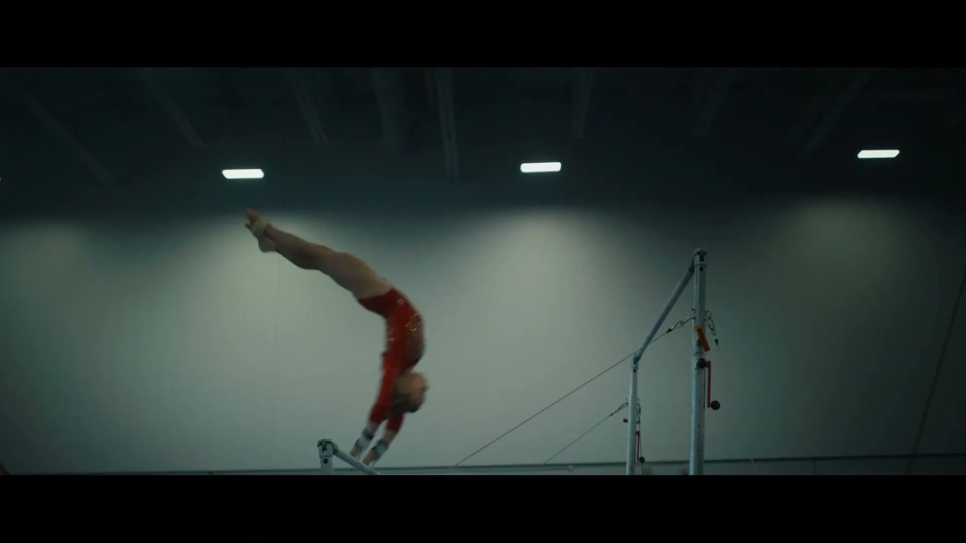A female gymnast in a red leotard performs a routine on the uneven bars in a brightly lit gymnasium.