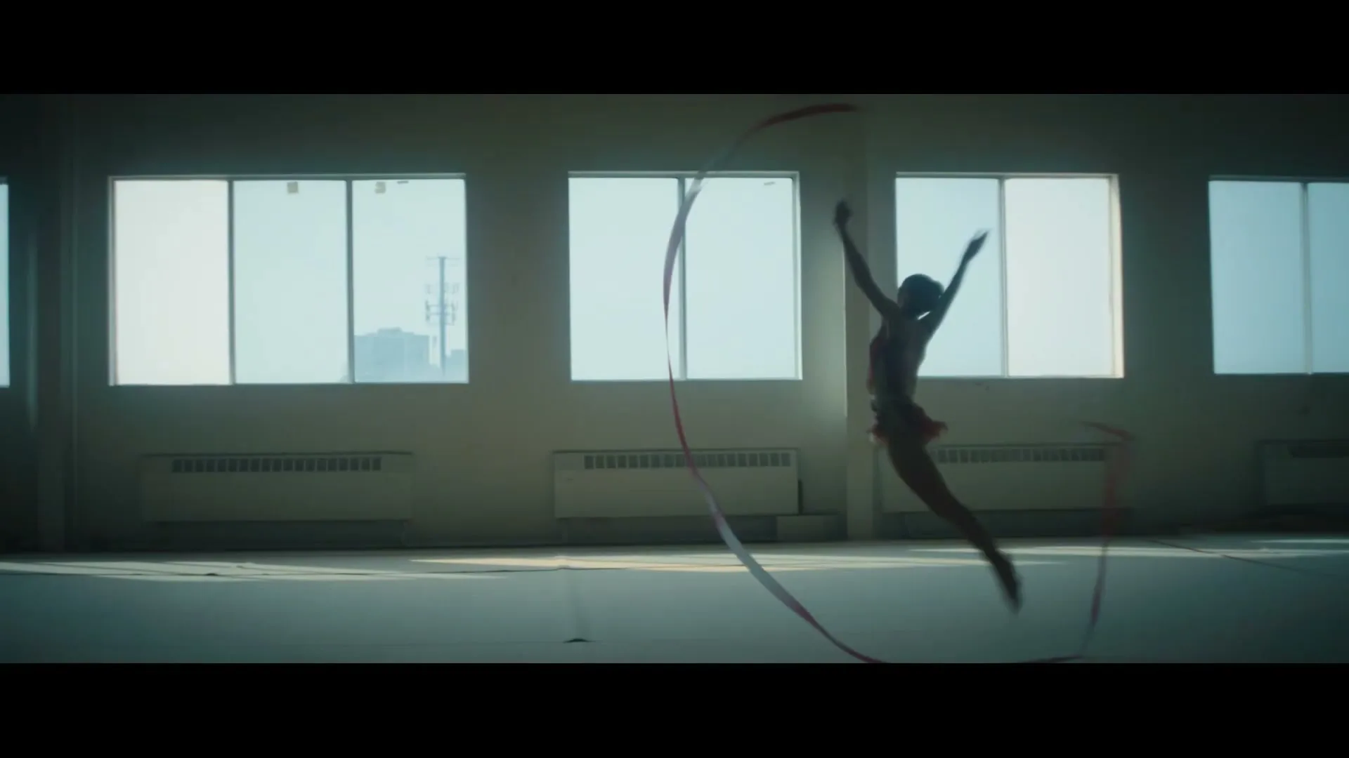 A female rhythmic gymnast in a leotard performs with a ribbon in a brightly lit gym with large windows.