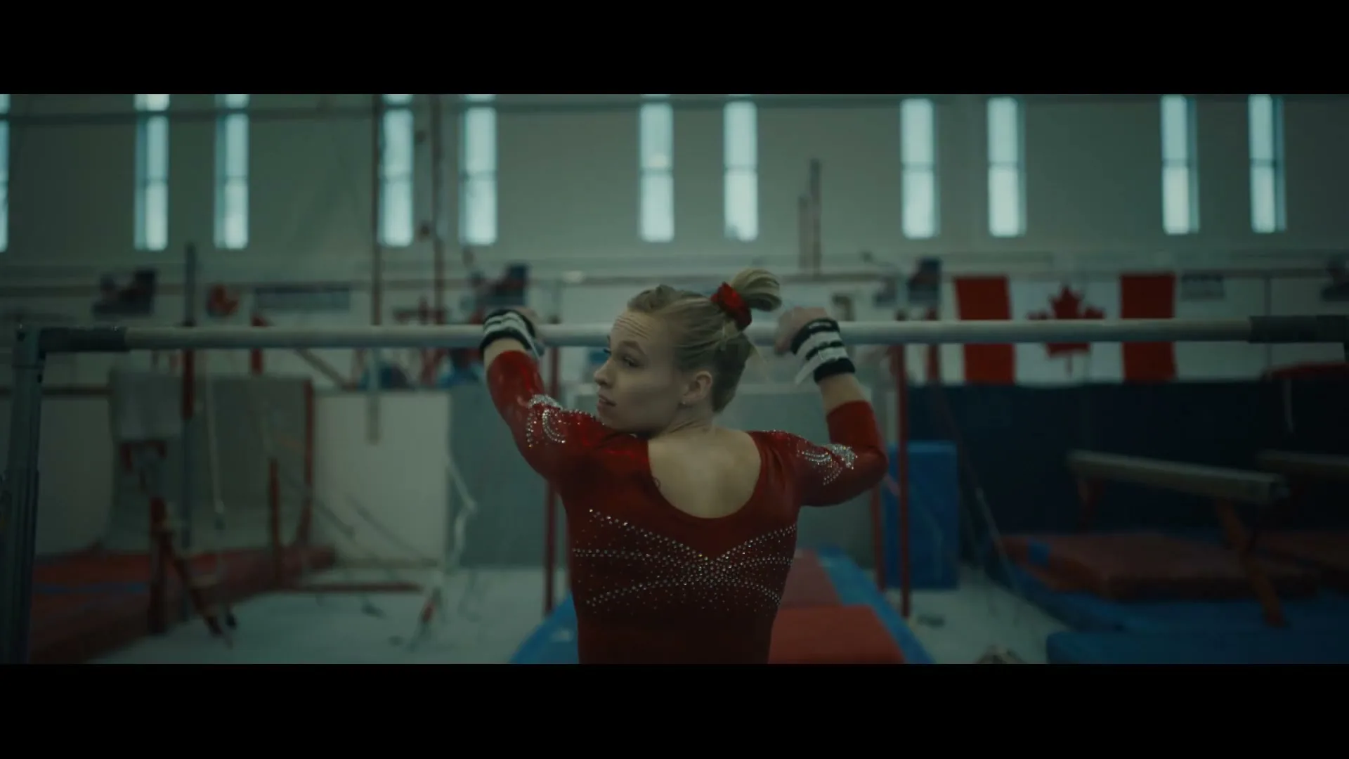 A female gymnast in a red leotard with a red bow in her hair, hangs on a bar in a gymnasium, looking over her shoulder.