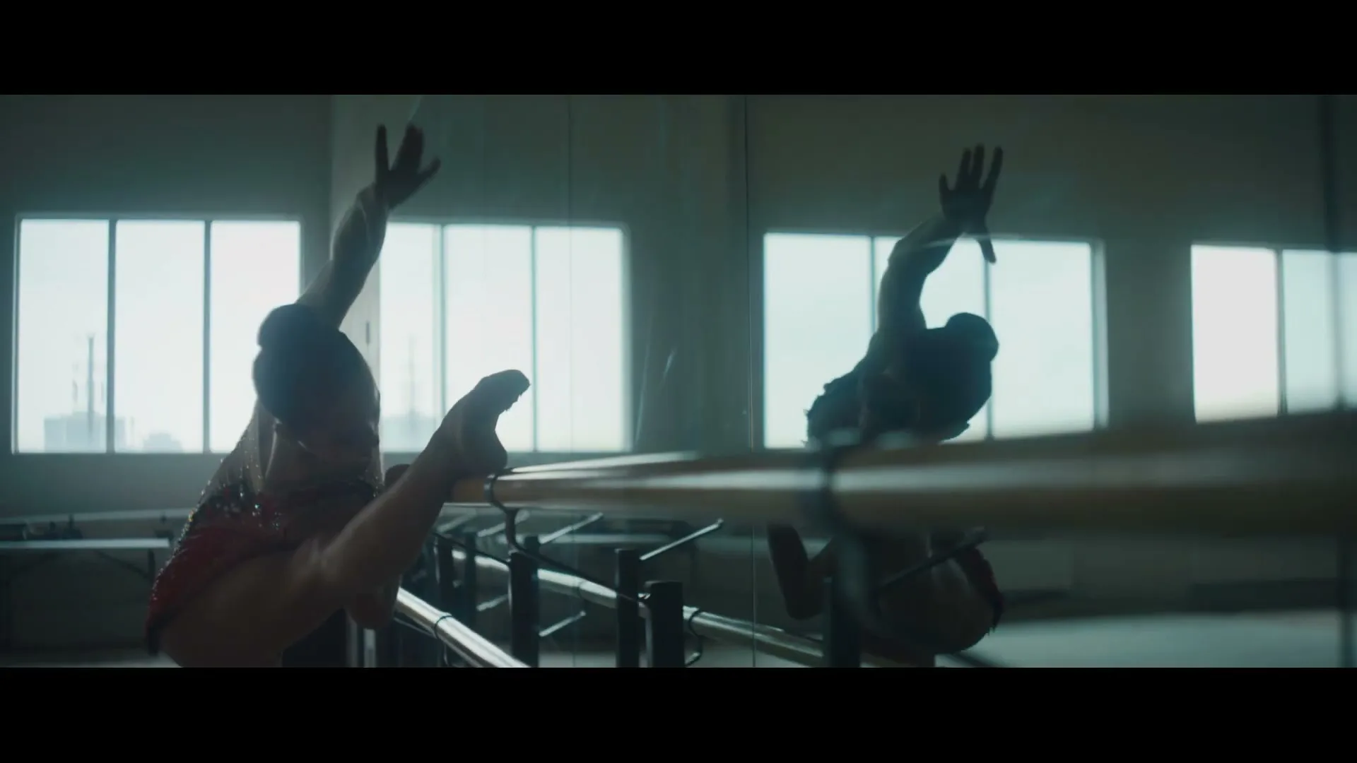 A female gymnast in a red leotard stretches at a ballet barre in a gym with large windows.