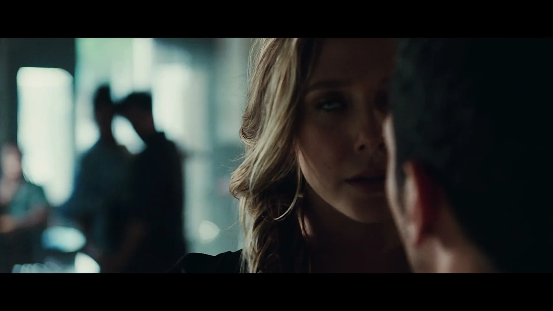 A woman with long, wavy hair looks intently at a man, her face partially obscured by his shoulder, in a dimly lit, crowded room.