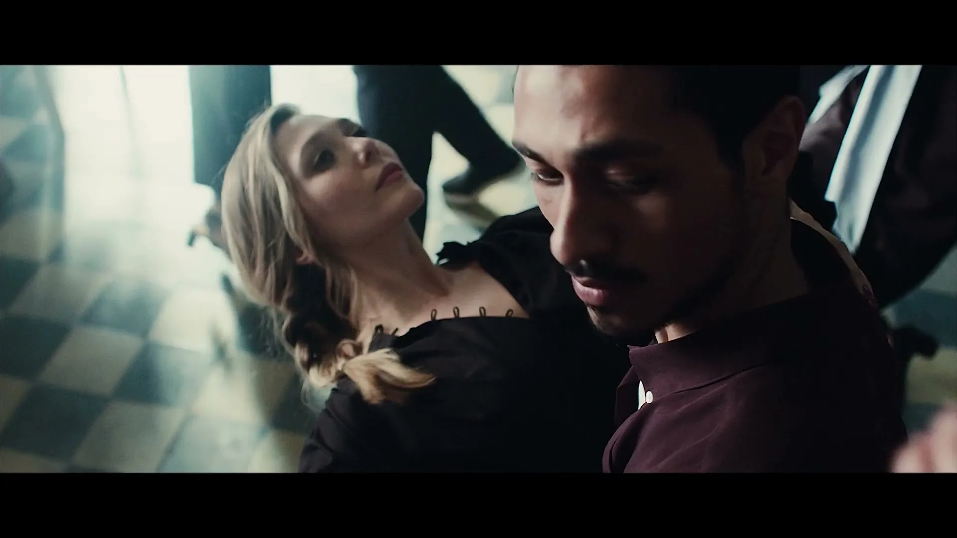 A man with a mustache holds a woman in a black dress as they dance in a room with a checkered floor.