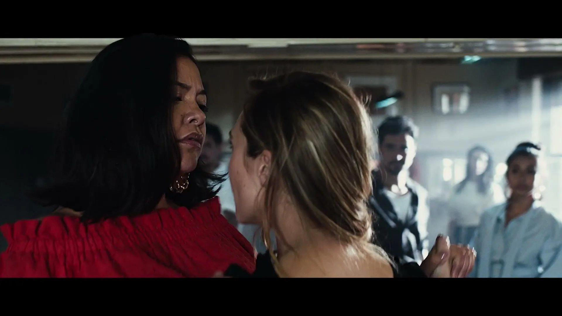 Two women dance closely, one in a red top and the other with her back to the camera, while other people are visible in the background of a dimly lit room.