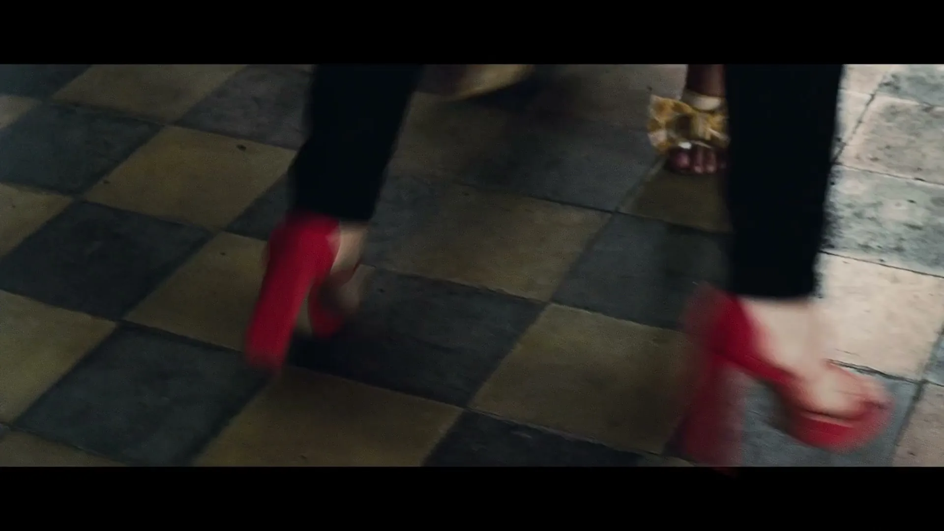 A woman in black pants and red high heels walks across a checkered floor, with another person's feet visible behind her.