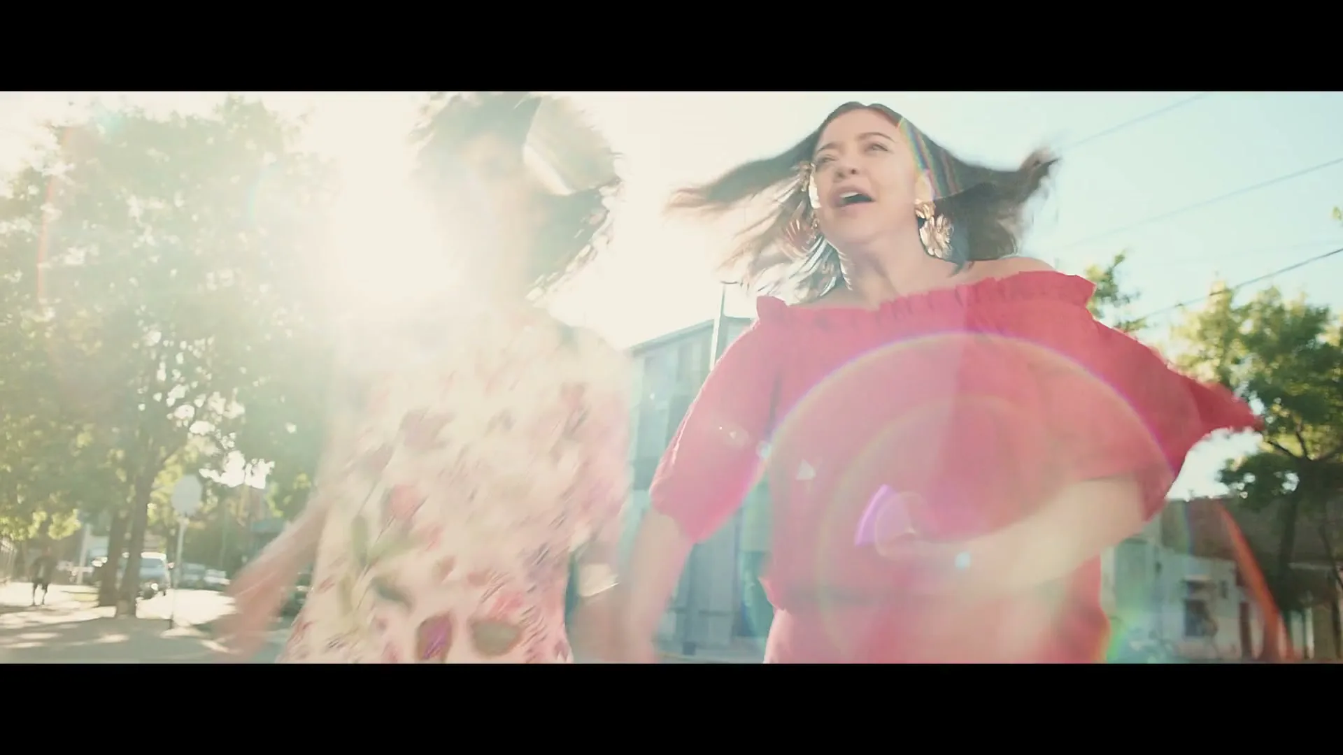 Two women, one in a red dress and one in a floral dress, dance joyfully on a sunny street with buildings and trees in the background.