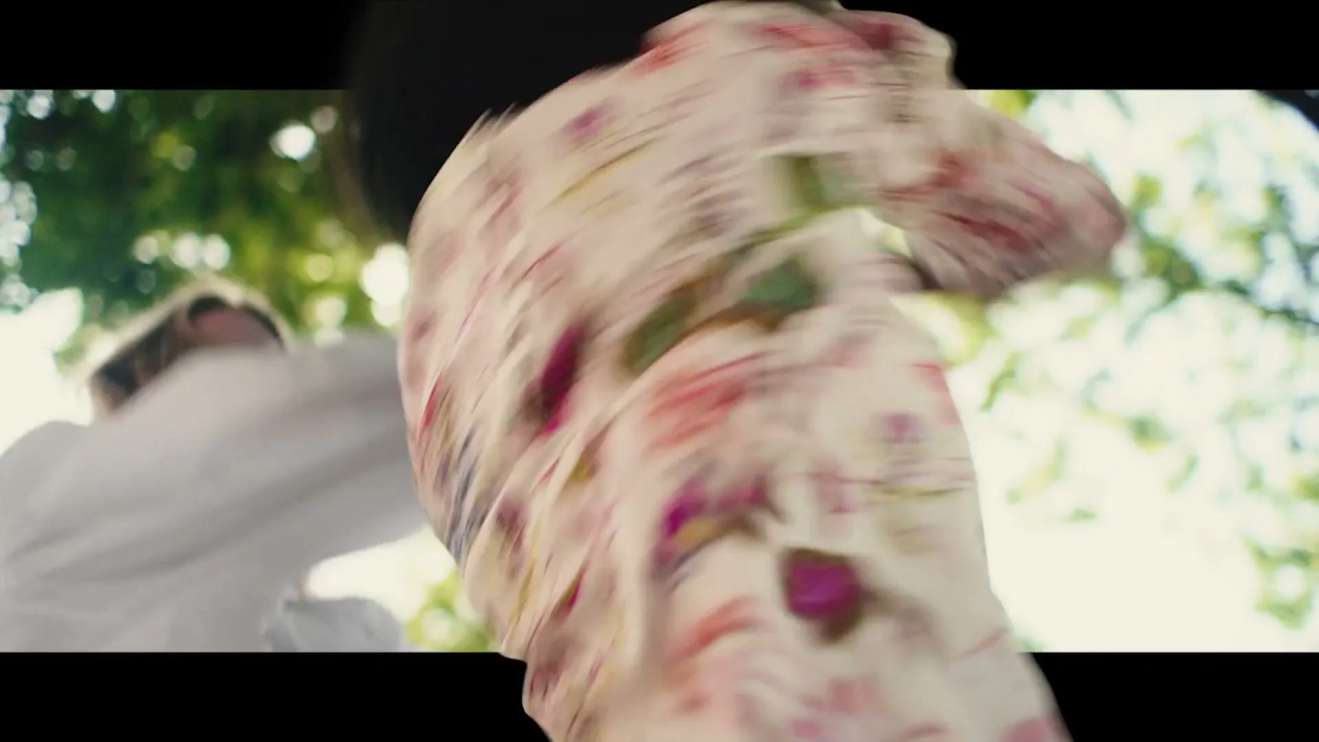 Two young women, one with curly dark hair and a floral top, the other with blonde hair and a white top, dance energetically outdoors under trees.