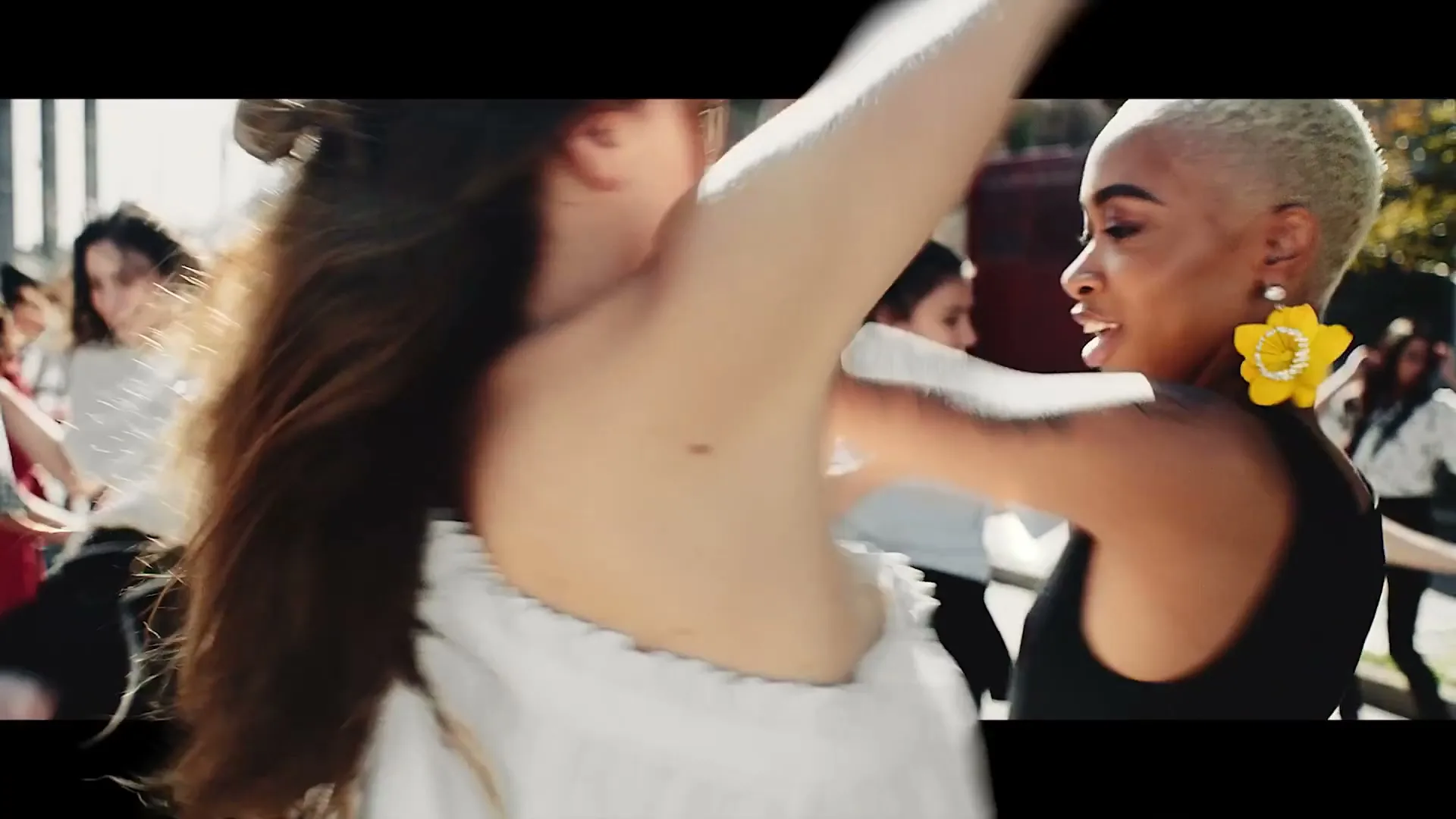 Two women dance closely, one with long dark hair in a white top, the other with short blonde hair and large yellow earrings, in an outdoor setting with other dancers.