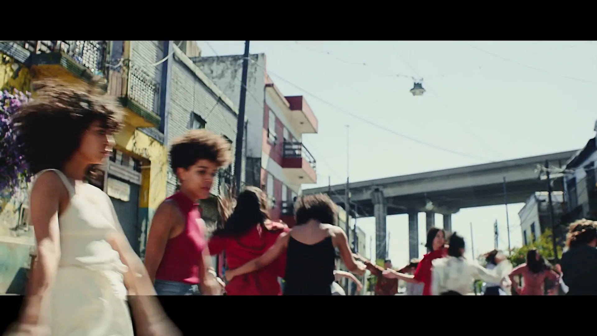 A diverse group of young people, mostly women, dance joyfully in a street with colorful buildings and an overpass in the background.