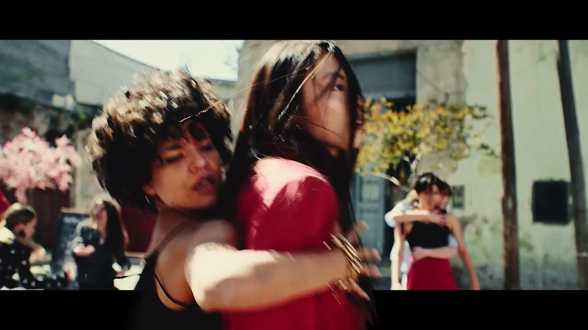 Two women dance closely, one in a red jacket and the other with curly hair, surrounded by other dancers in a sunny, urban street.