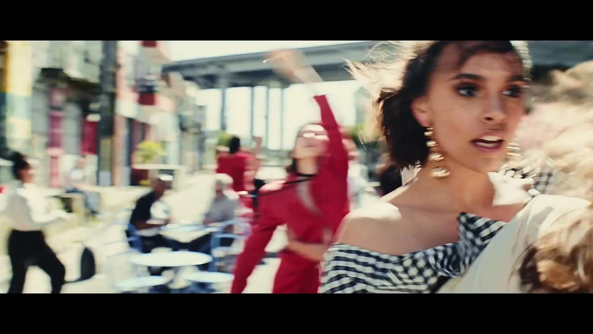 A young woman with dark curly hair and a gingham top dances energetically in a sunny outdoor cafe area, surrounded by other people.