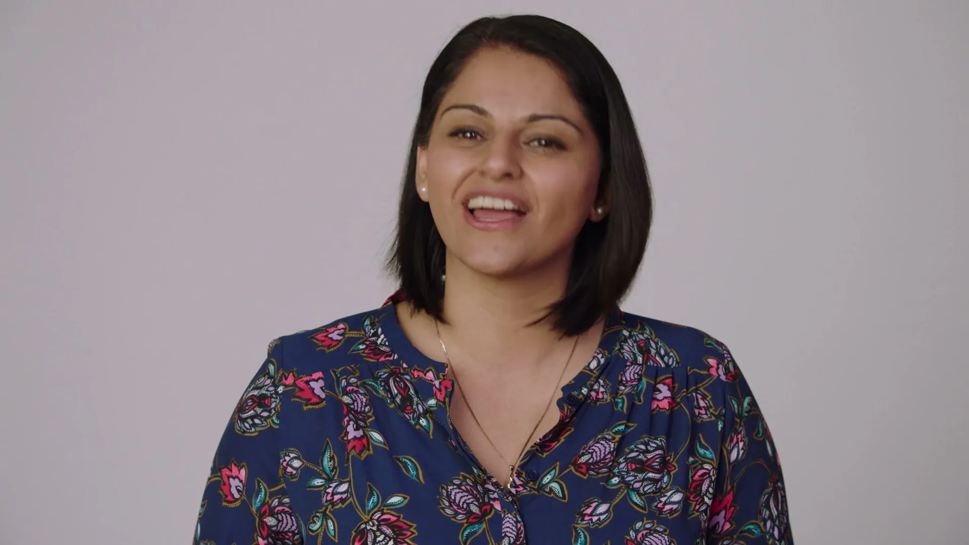 A woman with dark hair and a floral shirt speaks directly to the camera in a studio setting.