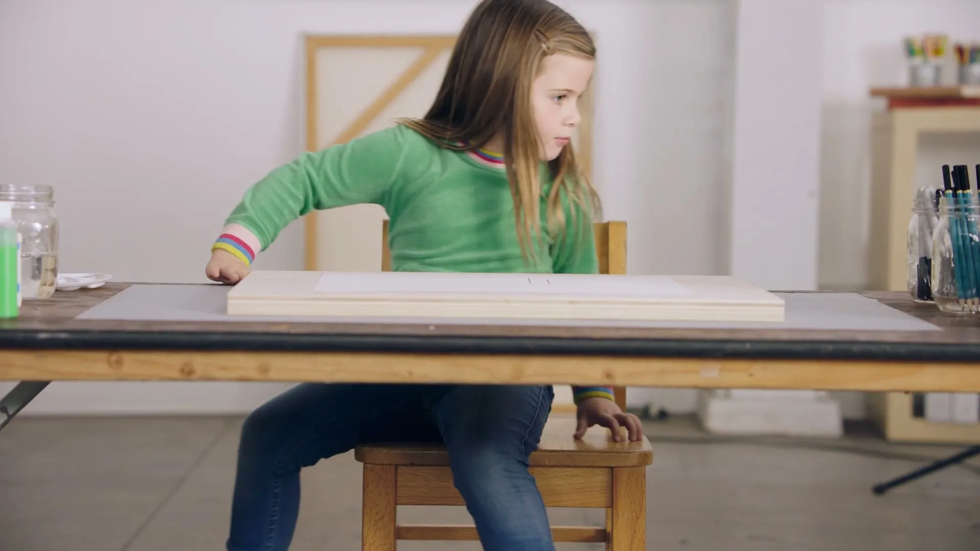 A young girl with long brown hair, wearing a green sweatshirt and jeans, sits at a wooden table in an art studio, looking to her right.