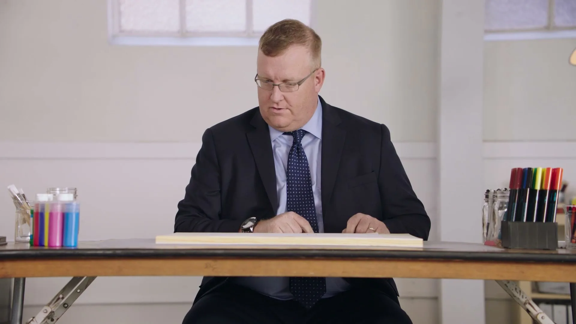 A man in his 40s, wearing a suit and glasses, sits at a wooden table, looking down at a blank piece of wood, appearing thoughtful.