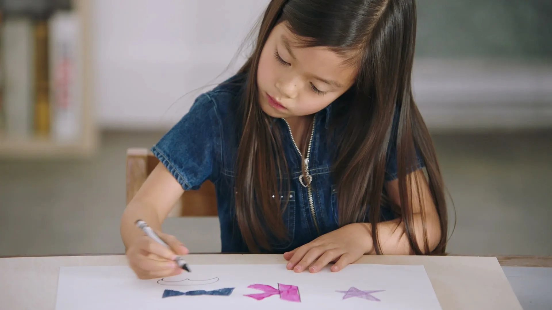 A young Asian girl with long dark hair, wearing a denim dress, sits at a table and draws on a piece of paper with a marker.
