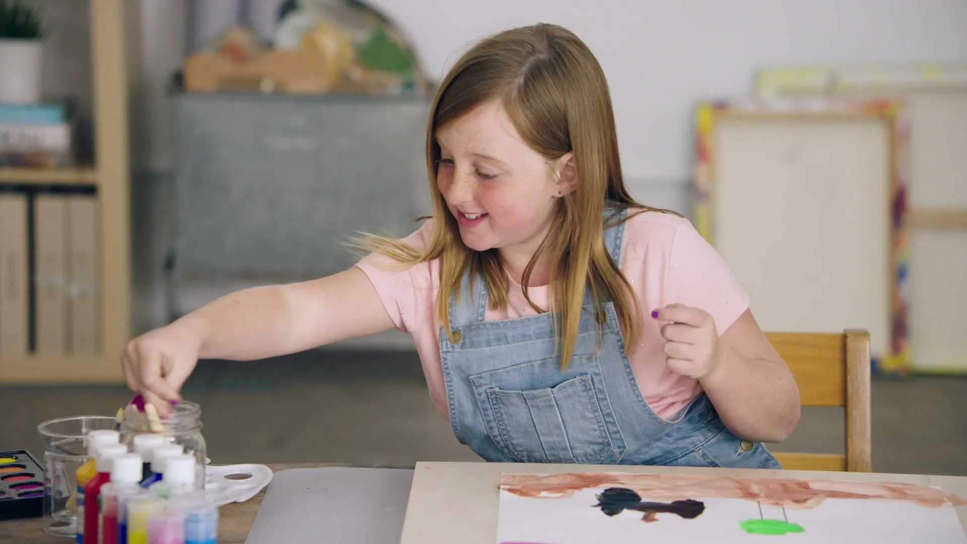 A young girl with long brown hair and freckles, wearing a pink shirt and denim overalls, smiles while painting at a table in an art studio.