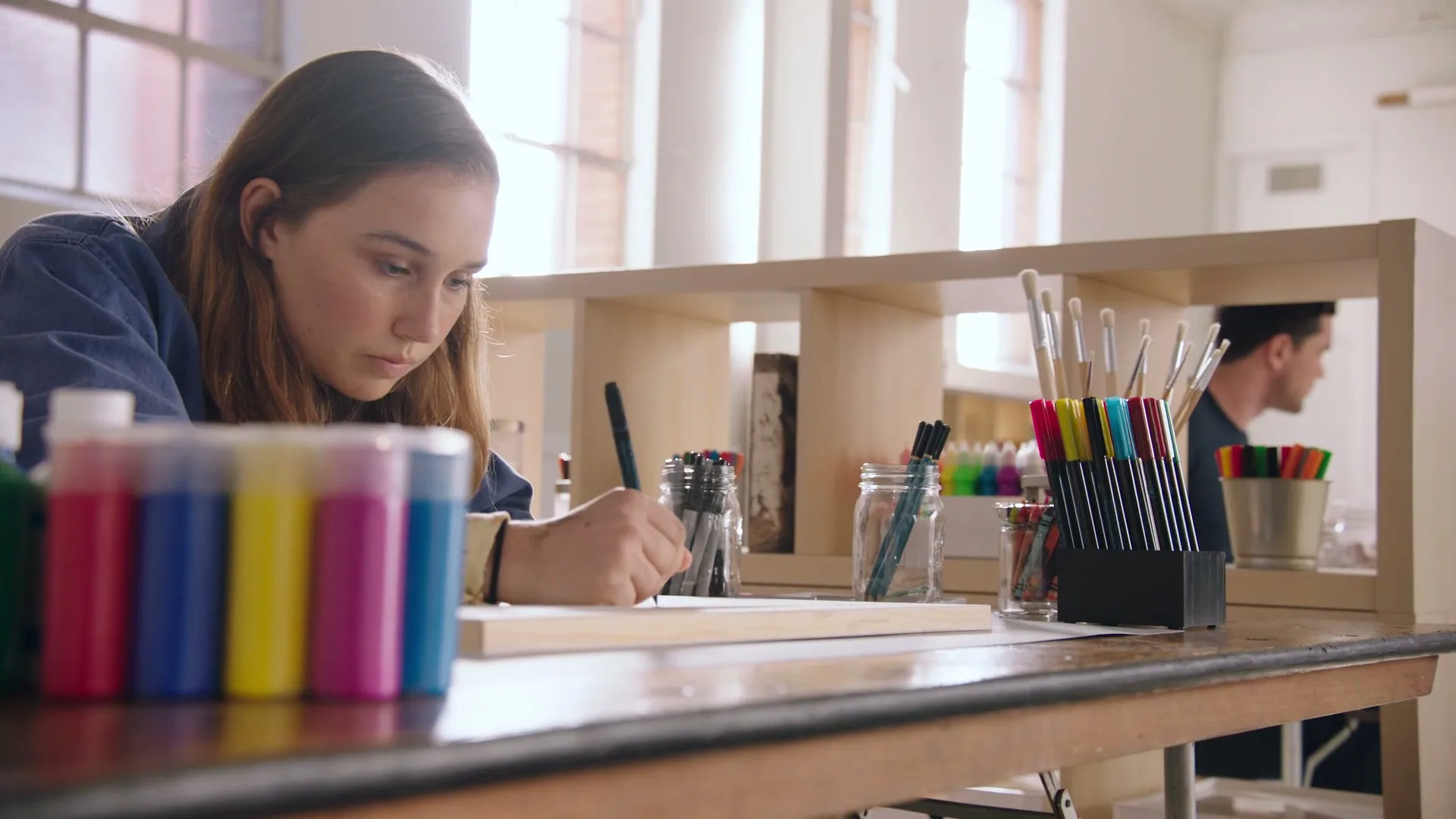 A young female artist with long brown hair, wearing a blue denim shirt, draws intently on a piece of wood at a table in a bright art studio.