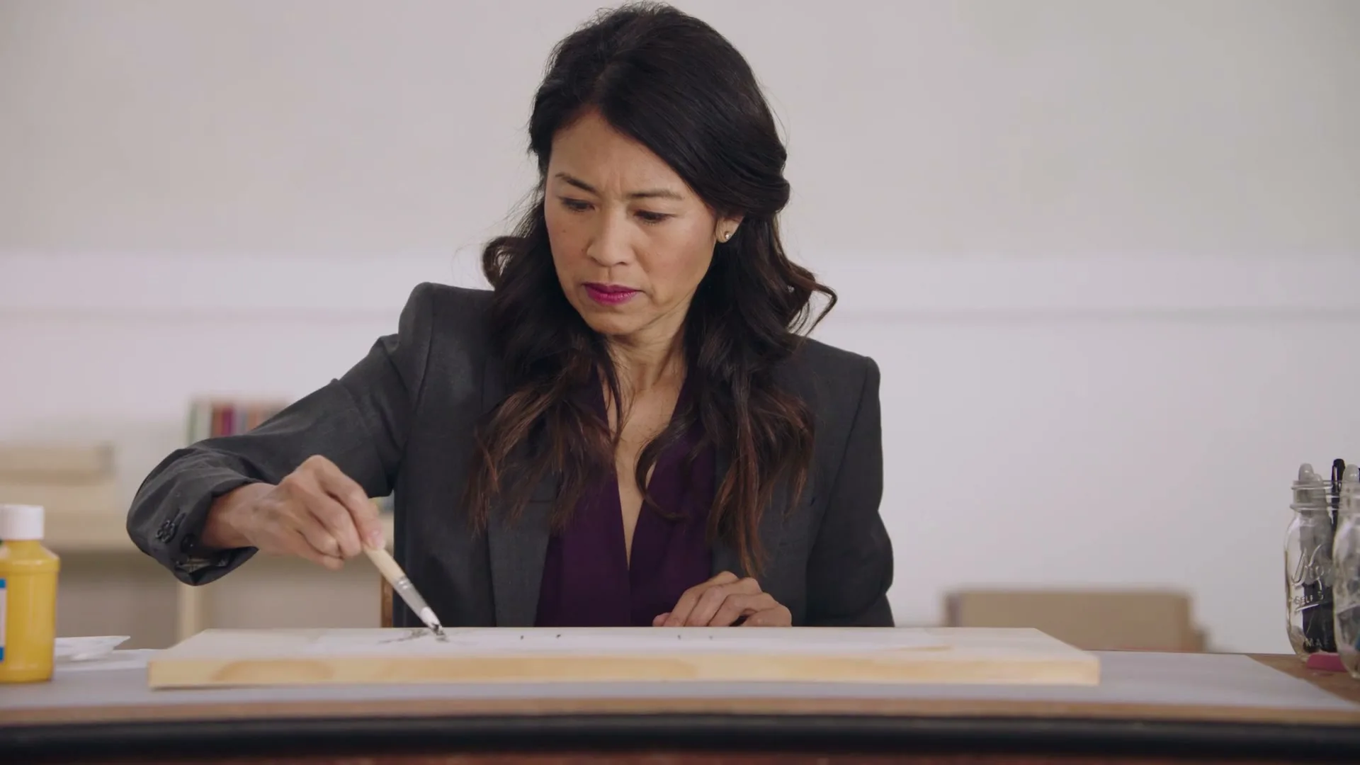 A focused Asian woman in her 40s, dressed in a business suit, meticulously paints on a wooden board with a small brush in a bright, minimalist interior.
