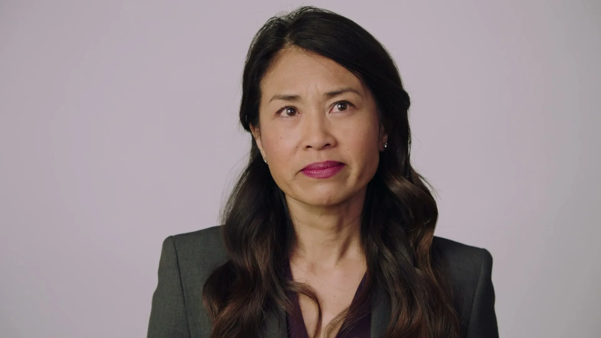 A woman with long dark hair and a dark suit speaks directly to the camera in a studio setting with a light background.