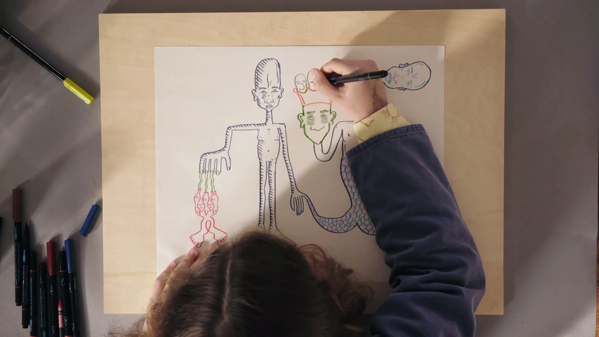 A child with brown hair and a blue jacket draws a whimsical, colorful creature on a piece of paper with a black marker, surrounded by other markers on a wooden board.
