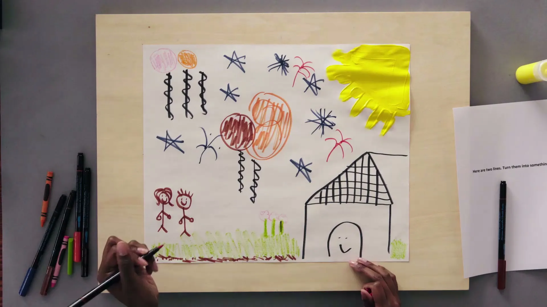 A child's hands are seen from an overhead shot, drawing on a piece of paper with crayons and markers, creating a colorful picture of a house, people, and balloons.