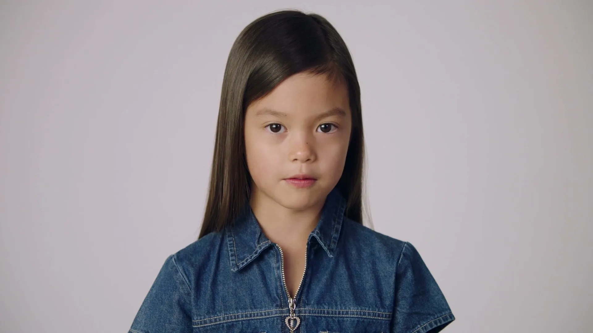 A young girl with long dark hair and a denim shirt speaks directly to the camera in a studio setting, expressing excitement.
