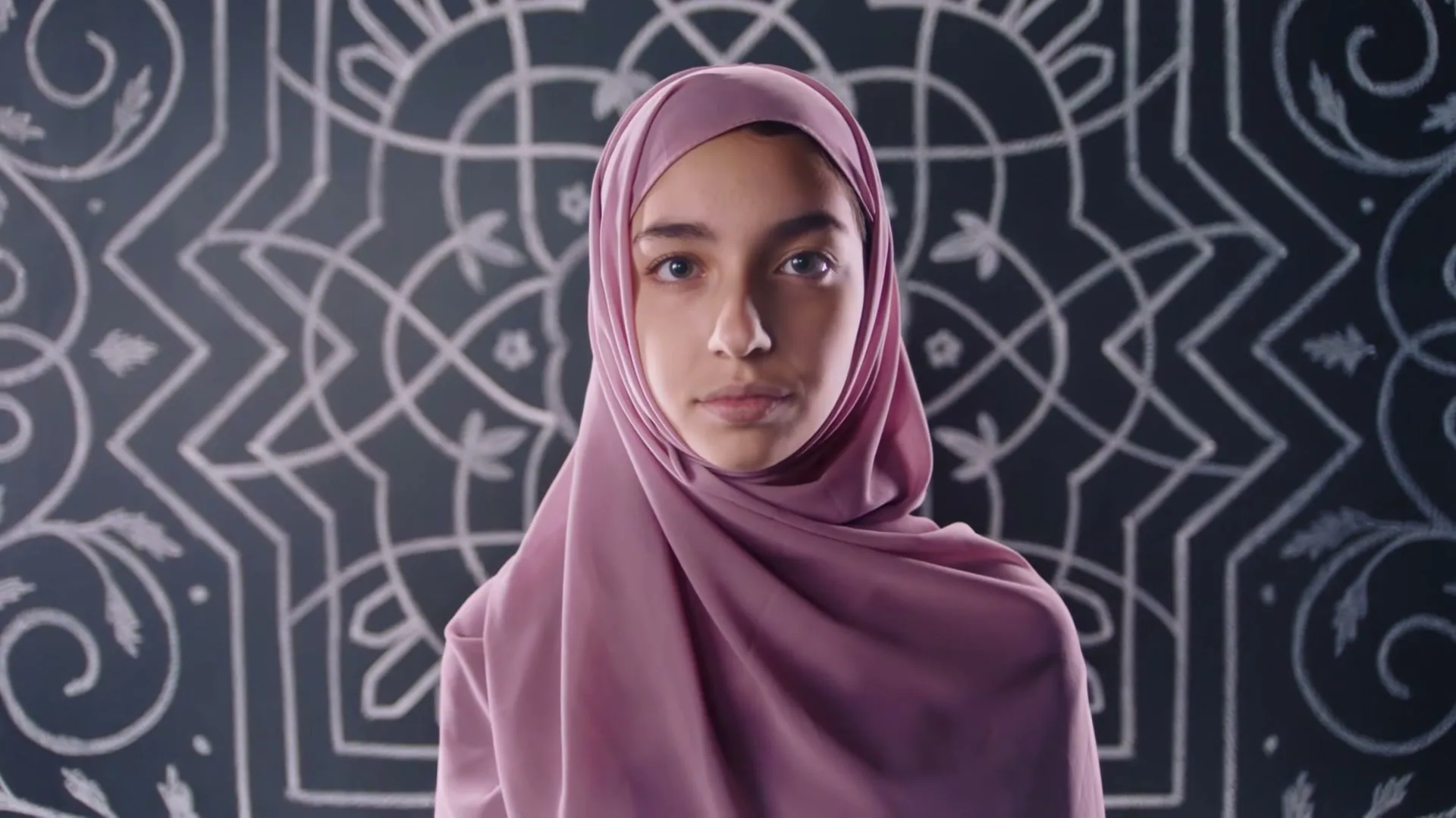 A young girl in a pink hijab stands in front of a chalkboard with white chalk drawings, looking directly at the camera.
