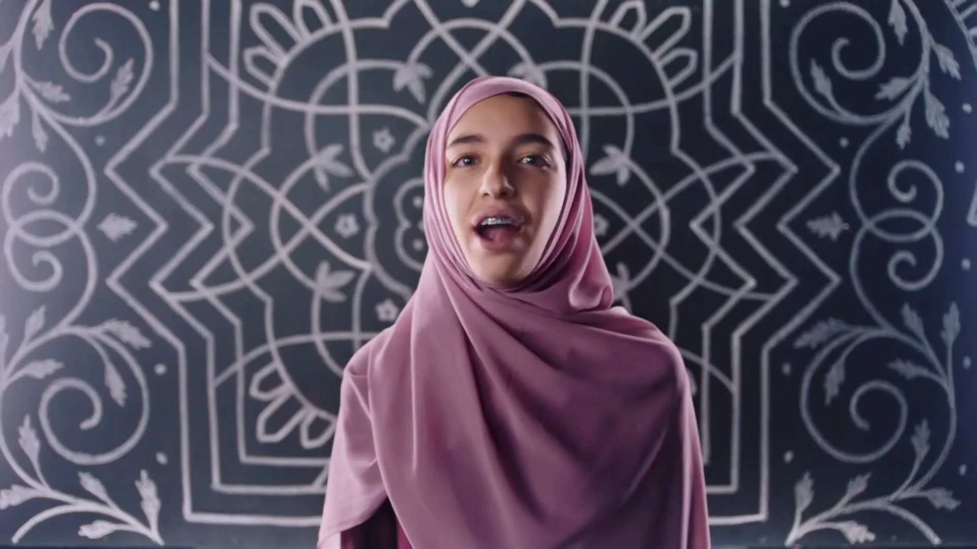 A young female in a pink hijab and braces speaks directly to the camera in front of a chalkboard with white chalk designs.