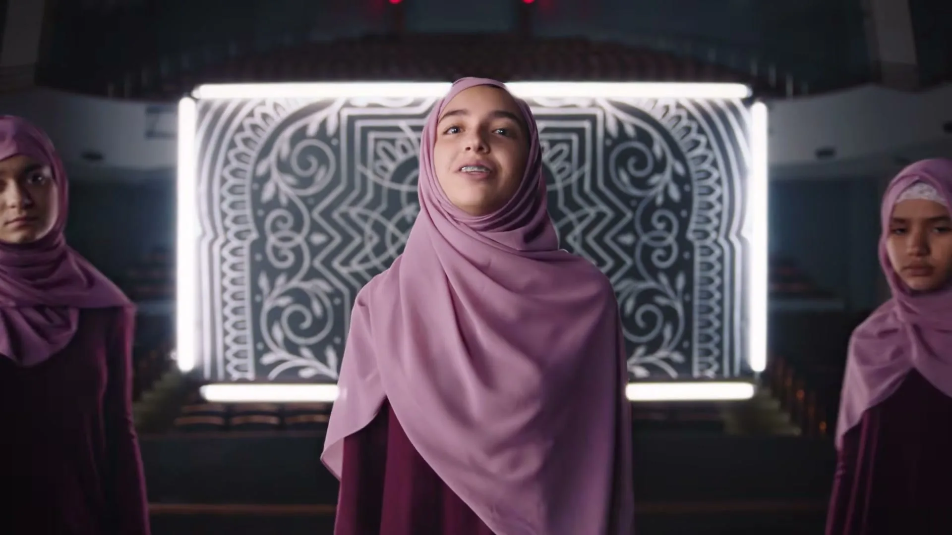 Three young girls in hijabs and maroon dresses sing and dance on a stage in an auditorium, with a glowing geometric pattern behind them.