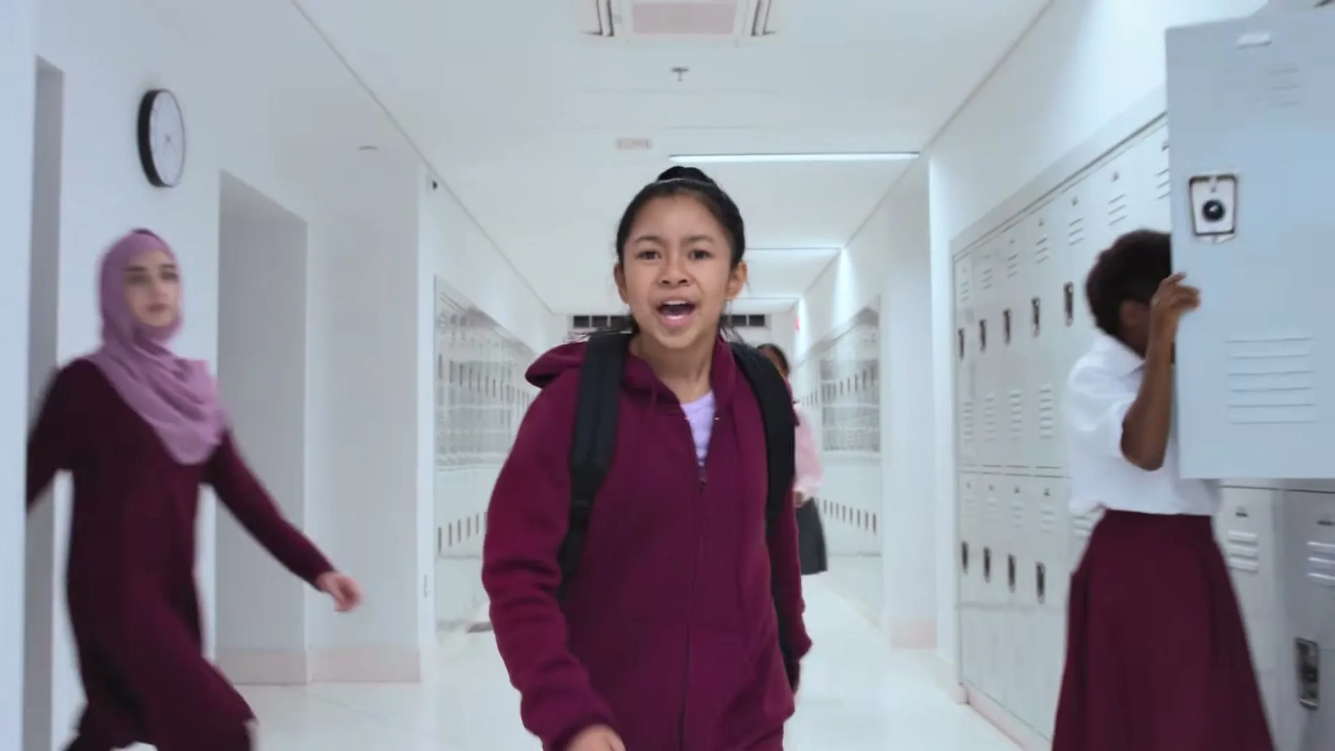 A young girl with a backpack walks down a bright school hallway, singing and gesturing, as other girls join her, dancing and smiling.