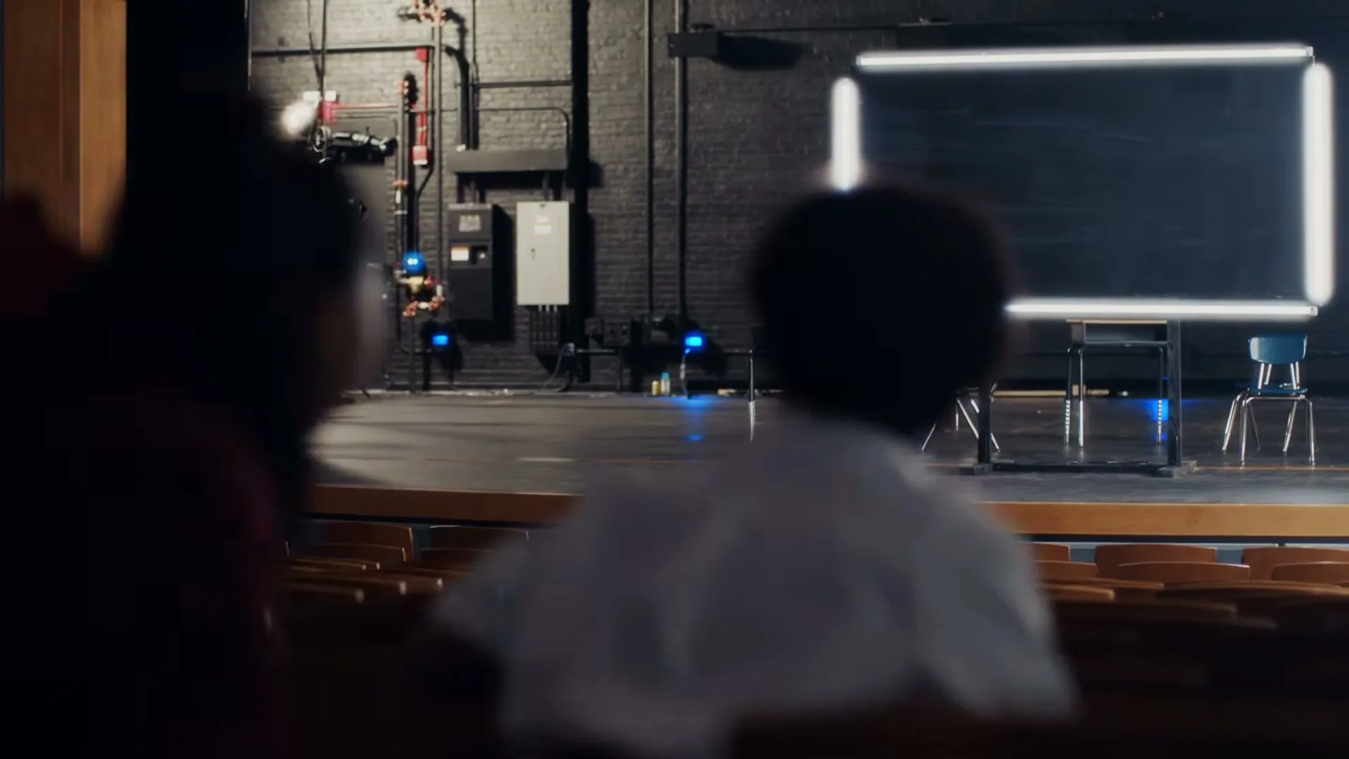A young girl with dark hair sits in an auditorium, looking towards a stage with a glowing rectangular screen and chairs, as other blurred figures sit in the foreground.