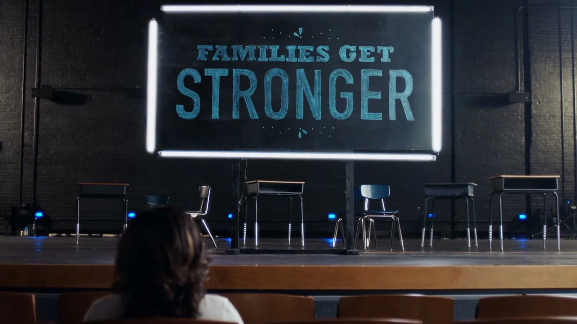 A woman with dark hair sits in an auditorium, looking at a stage with school desks and a large screen displaying animated text.