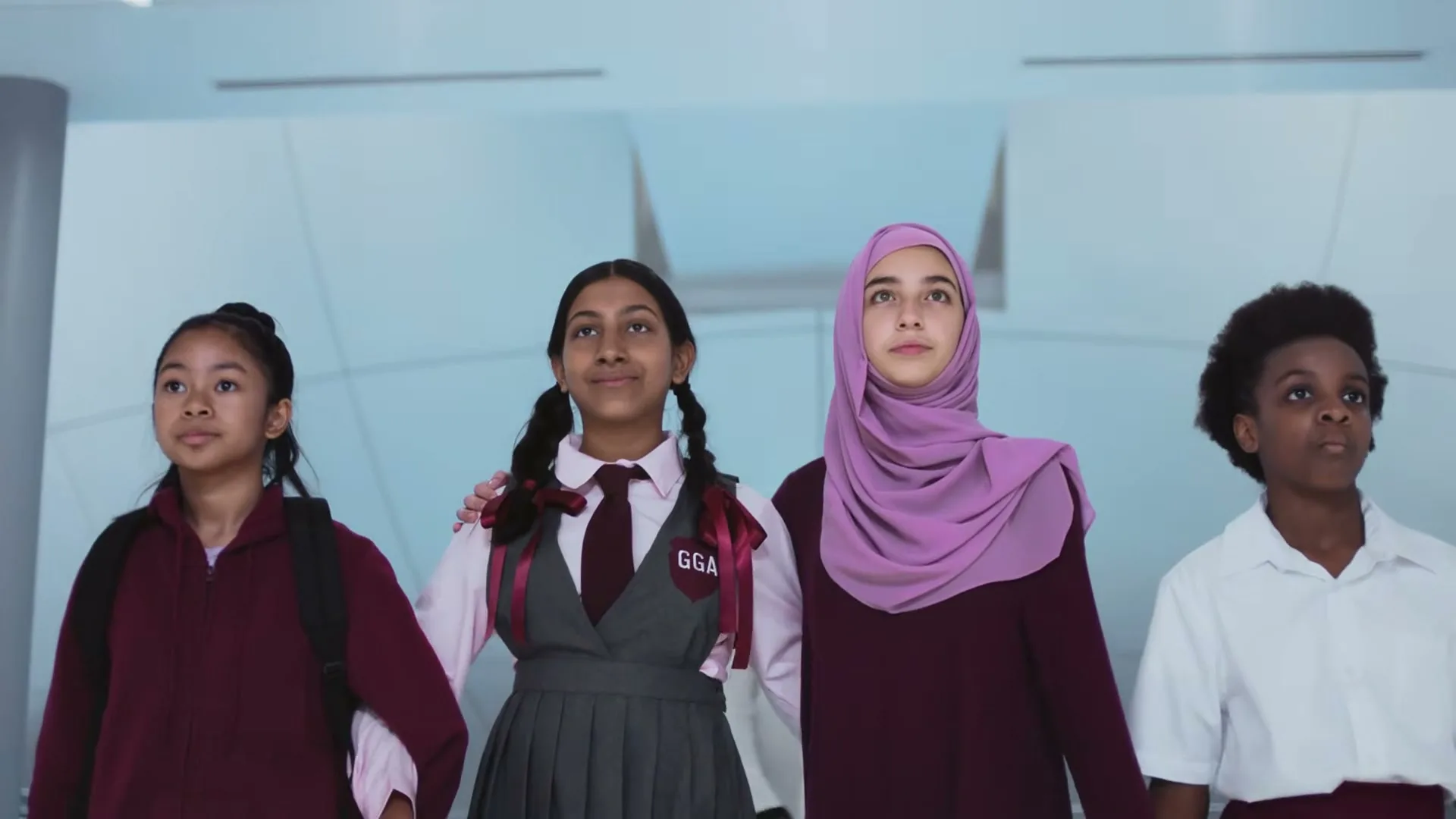 Four diverse schoolgirls, dressed in school uniforms and casual wear, stand together looking up with determined expressions in a modern, bright hallway.