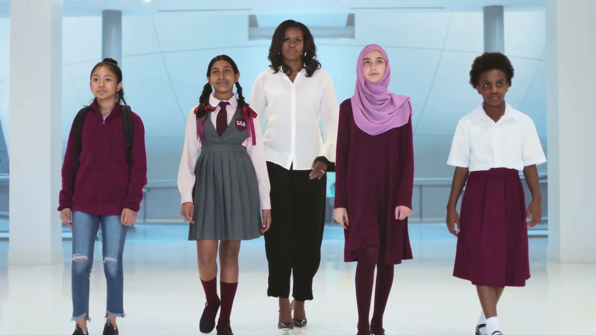Michelle Obama walks forward with four diverse young girls, all smiling, in a bright, modern, minimalist hallway.