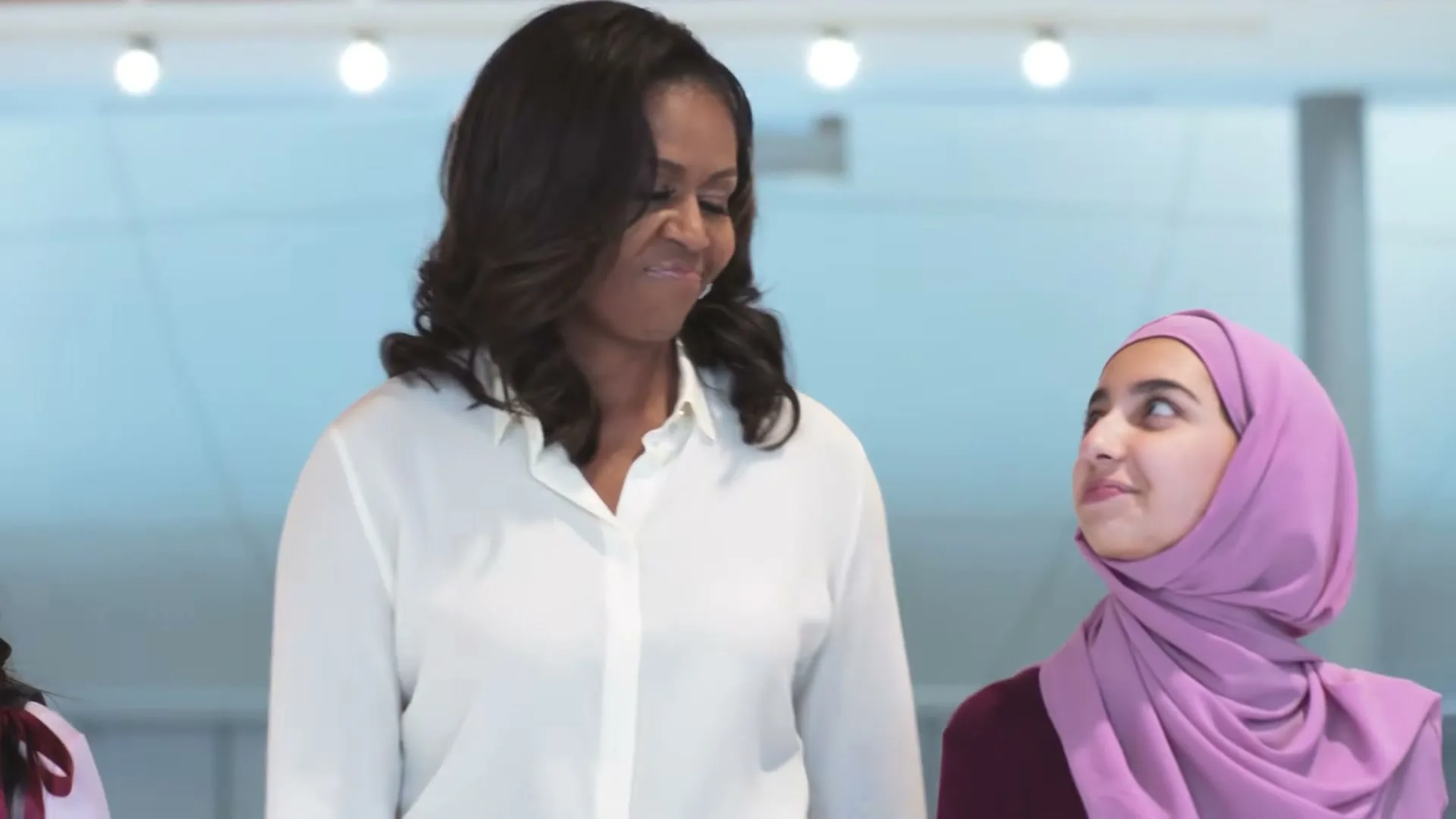 Michelle Obama stands between two young girls, one wearing a hijab, all smiling and looking forward in a bright, modern interior.
