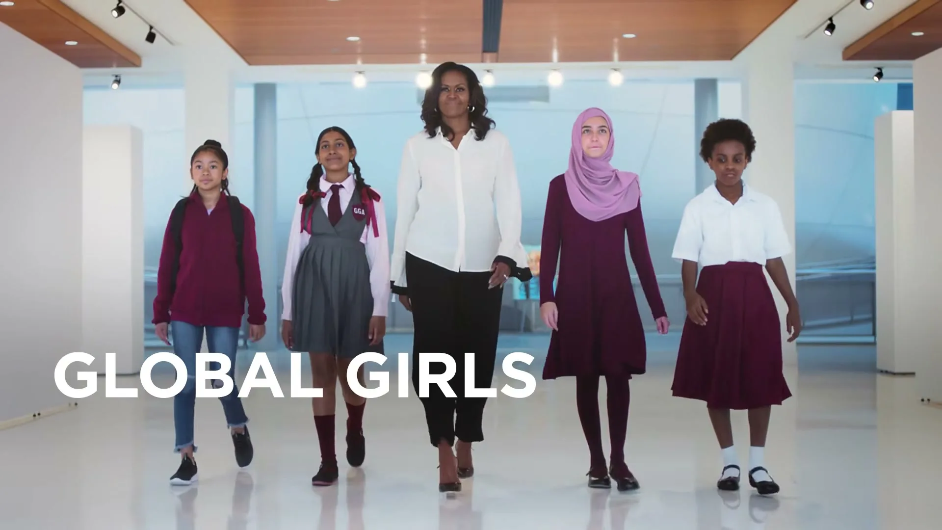 Michelle Obama walks forward with four diverse schoolgirls, all smiling, in a bright, modern interior space with white walls and a light wood ceiling.