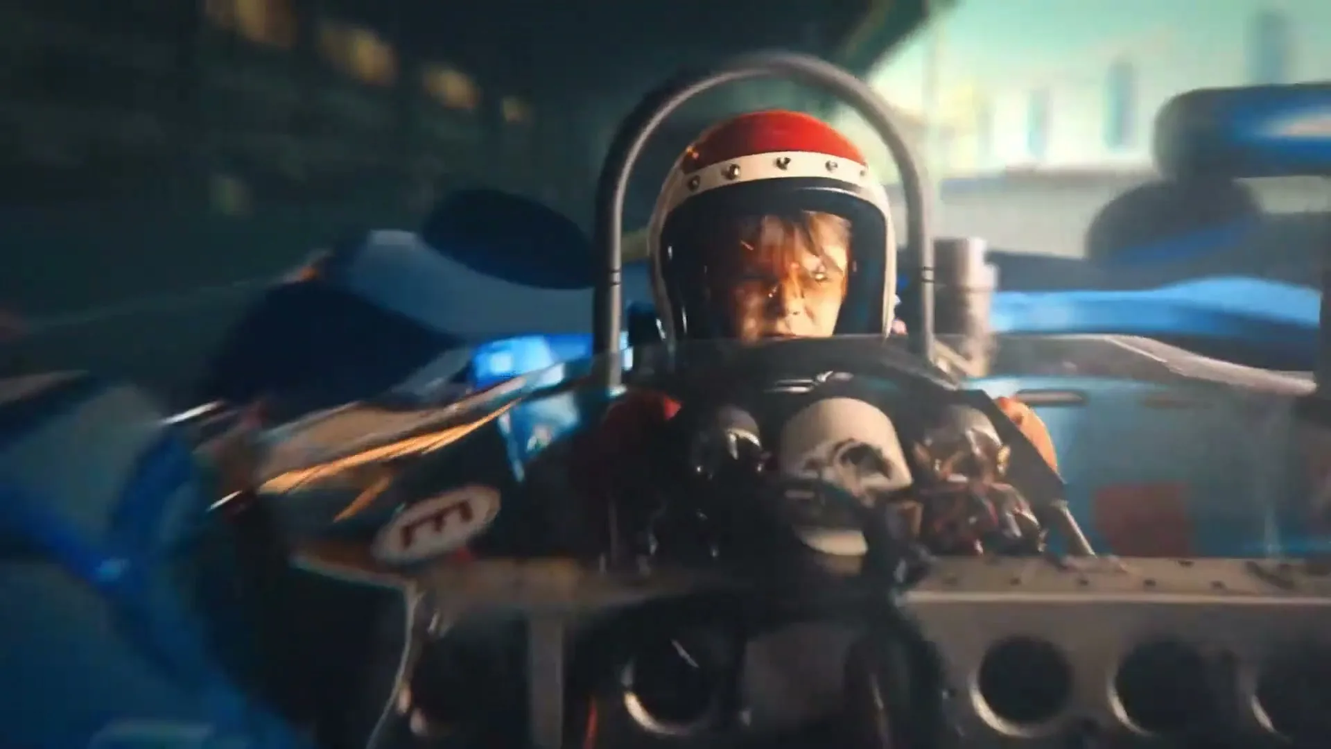 A young boy in a racing helmet and goggles drives a blue race car with the number 67 on it, looking determined on a race track.