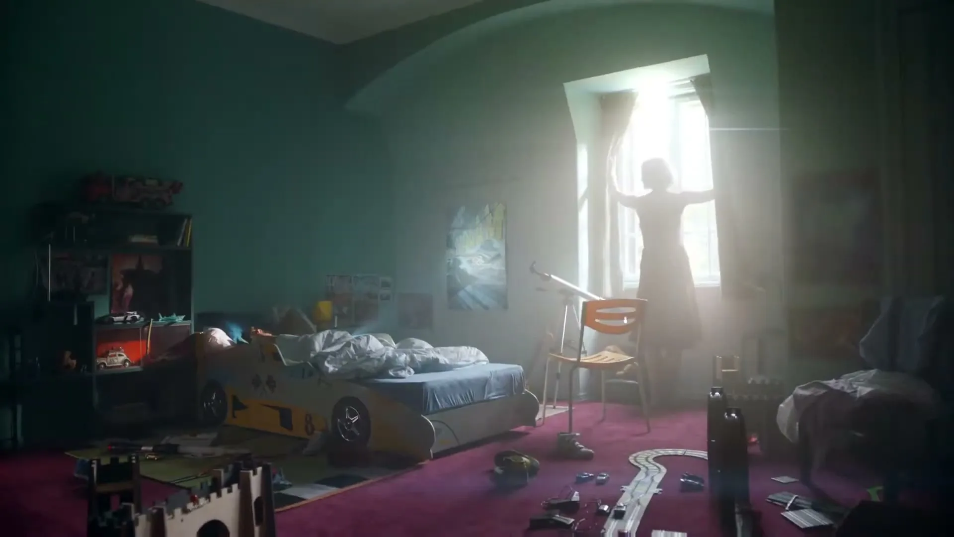 A child stands silhouetted against a bright window, opening curtains in a messy bedroom with a race car bed.
