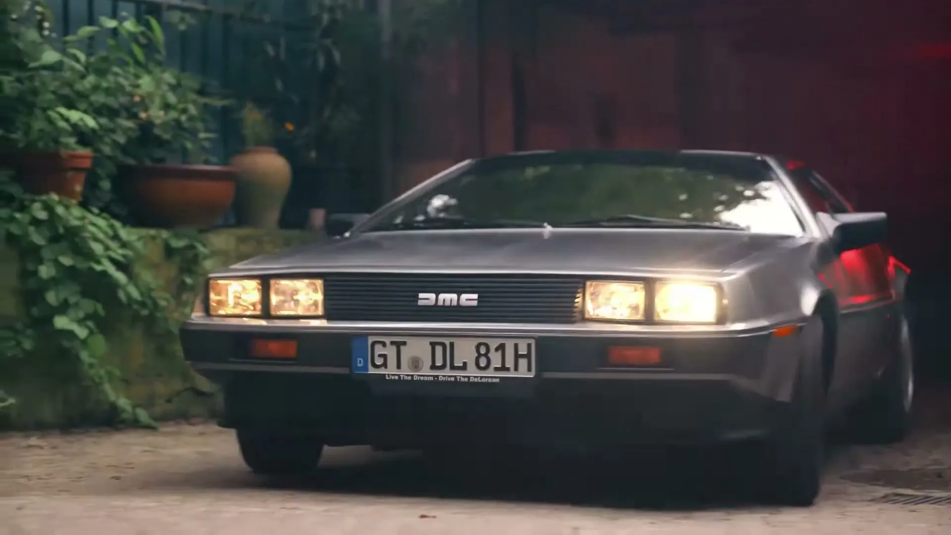 A silver DeLorean car with its headlights on slowly reverses out of a dark garage into a leafy exterior setting.