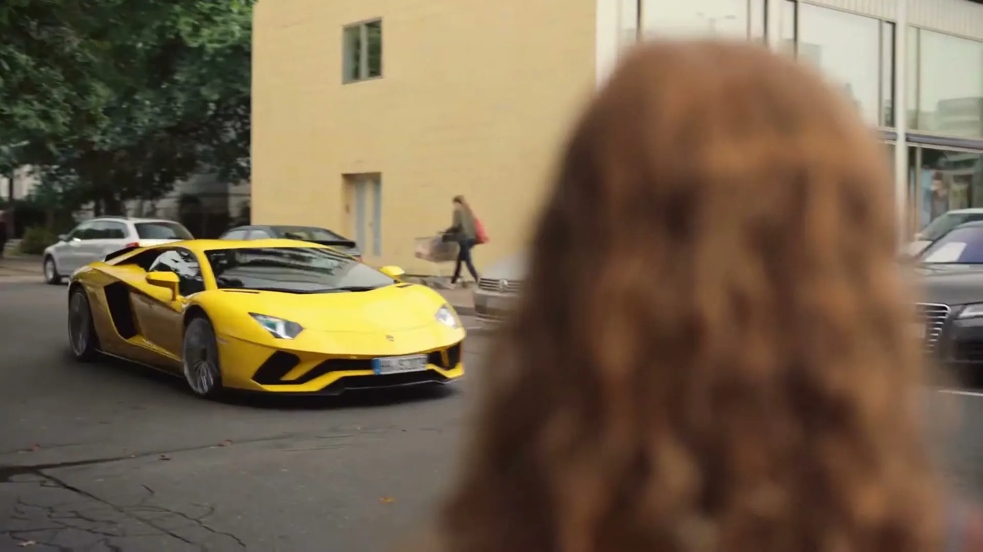A young girl with long, curly brown hair watches a bright yellow Lamborghini sports car drive past her on a city street during the day.