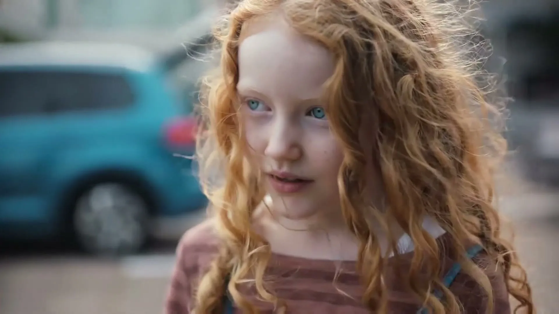 A young girl with long, curly red hair and blue eyes looks around with a curious expression, with a blue car in the background.