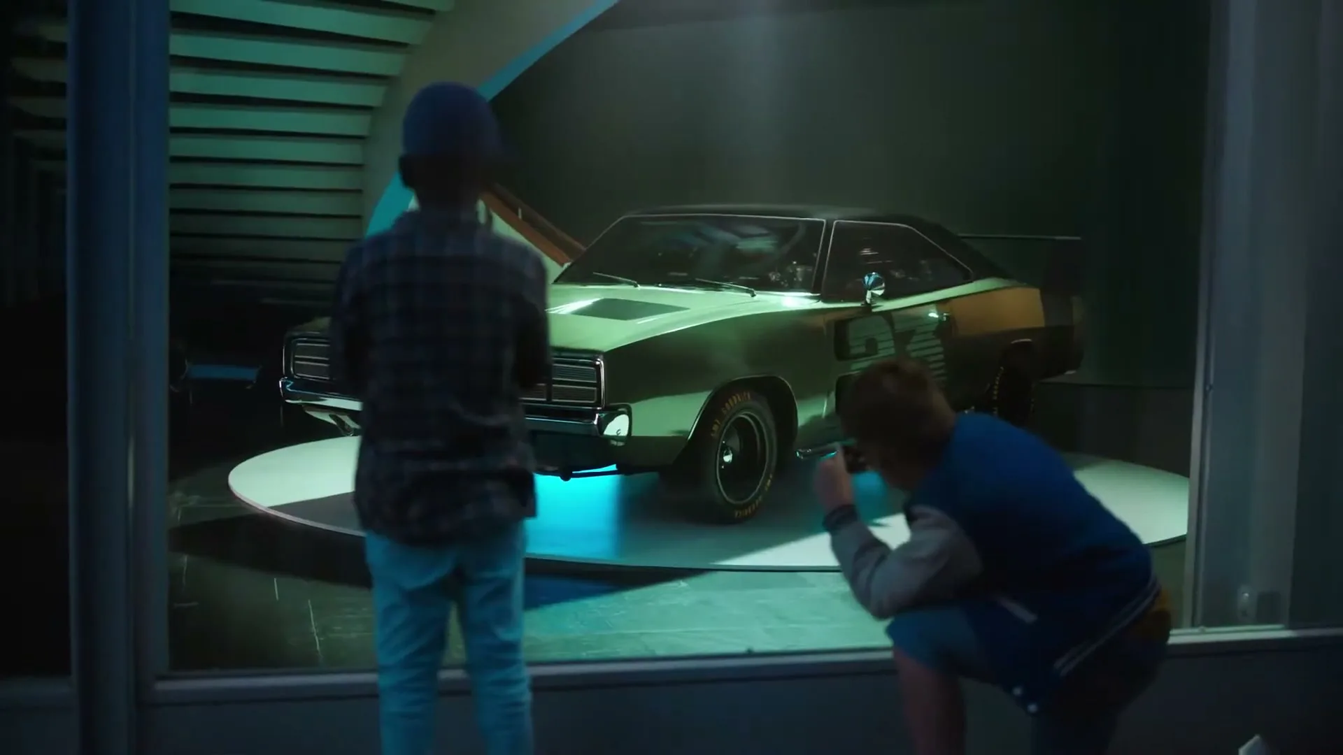 Two young boys look at a classic green muscle car on display in a showroom window at night, one boy takes a photo.