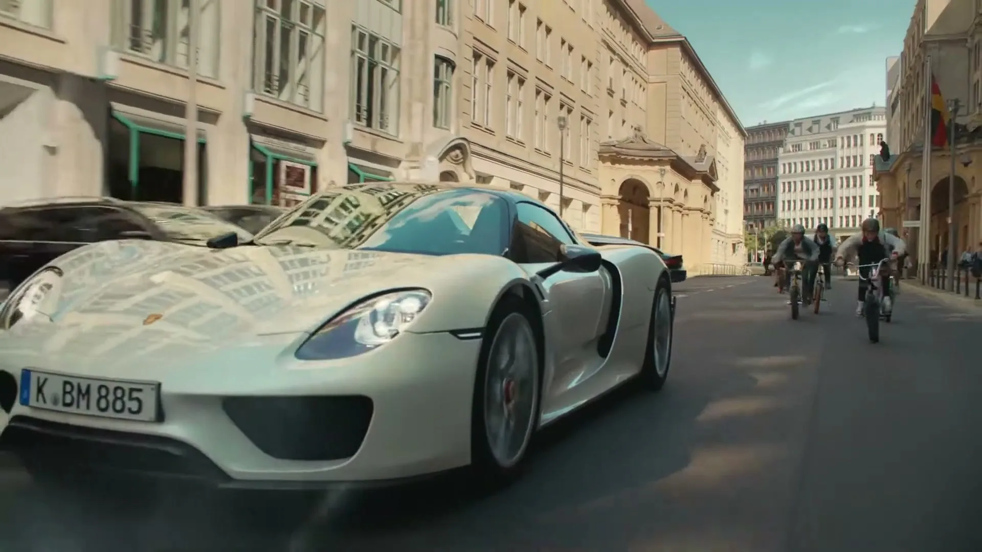 A white Porsche sports car drives down a city street, followed by a group of young men on BMX bikes, under a bright sky.