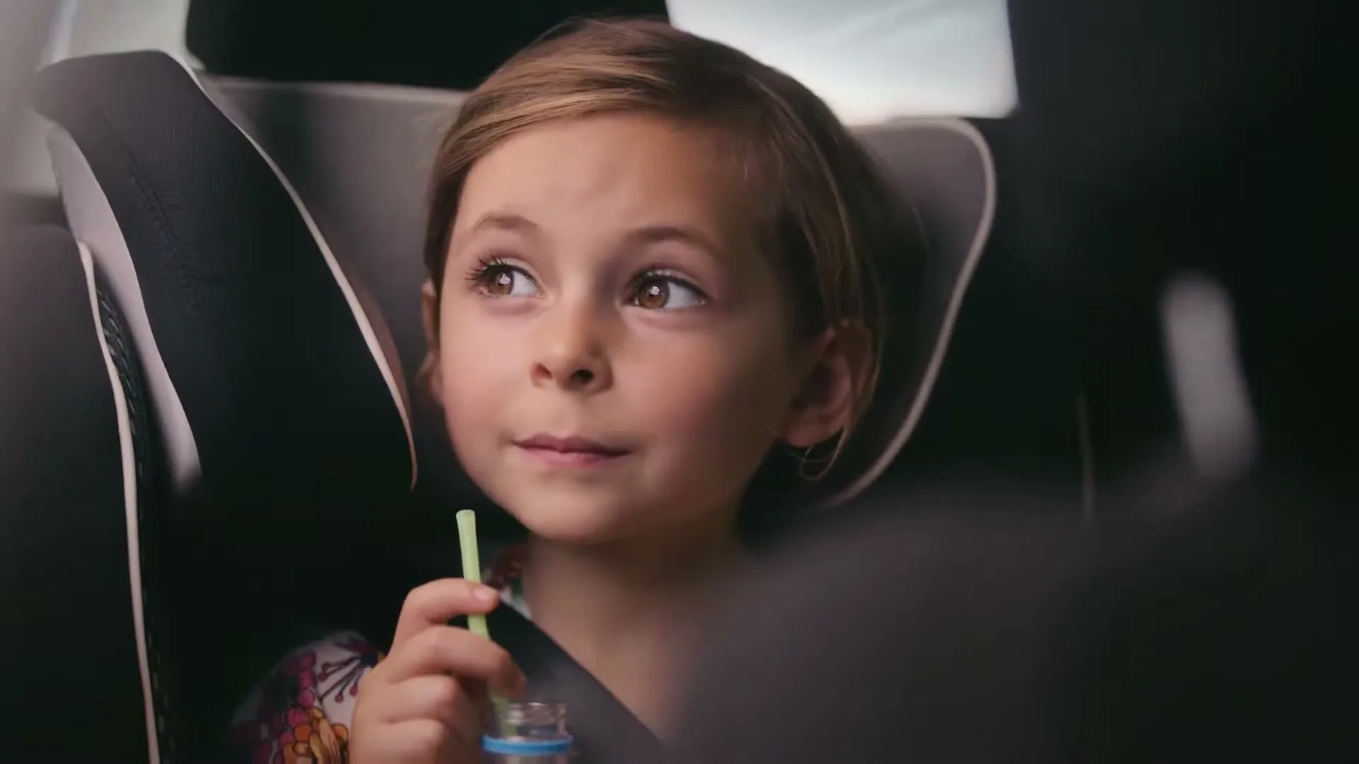 A young girl with light brown hair, wearing a colorful top and seatbelt, sips from a straw in a car seat, looking thoughtfully out the window.