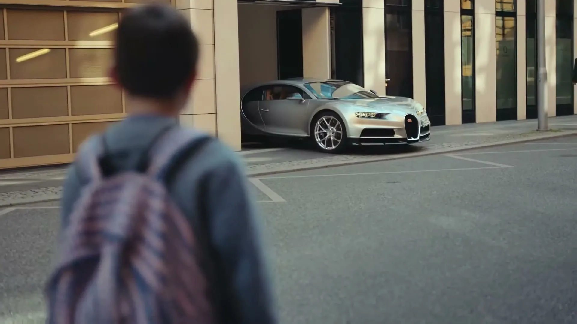 A young boy with a backpack walks away from the camera, looking at a silver sports car emerging from a garage on a city street.