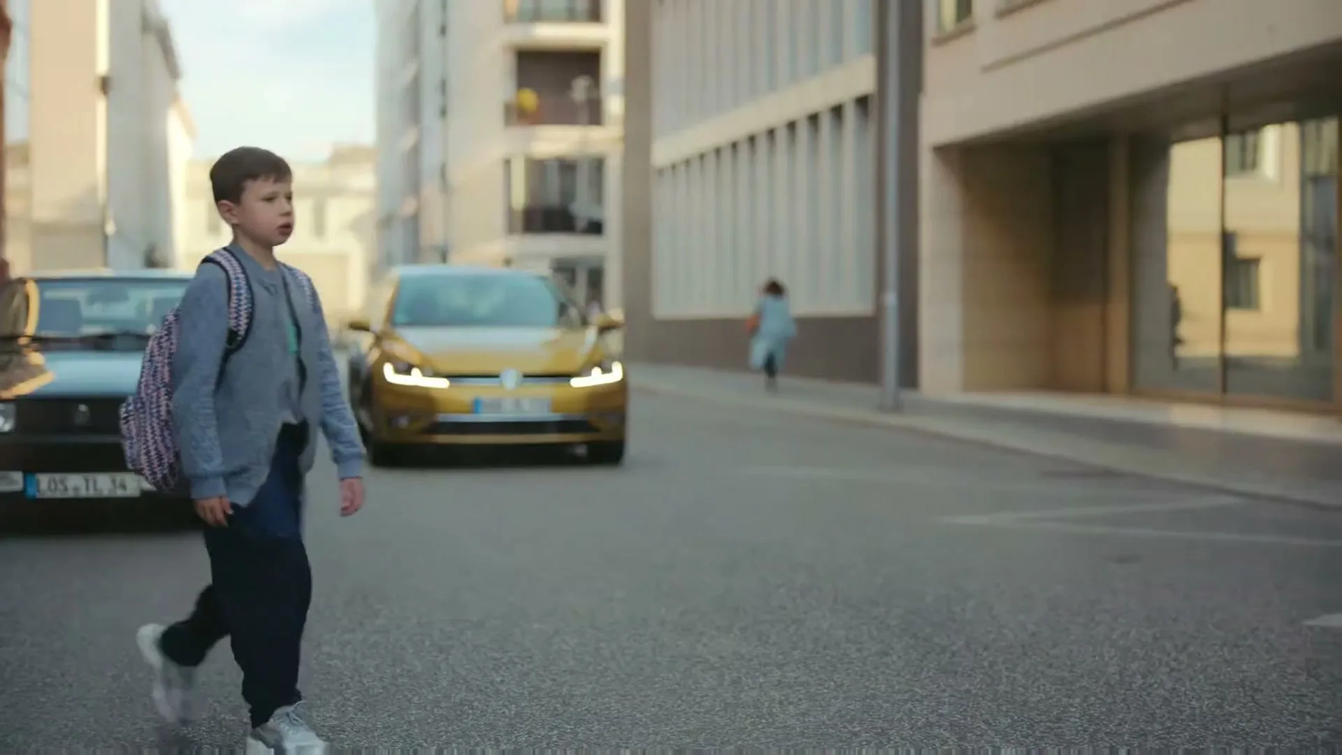 A young boy with a backpack crosses a city street, looking left as a gold car approaches in the background.